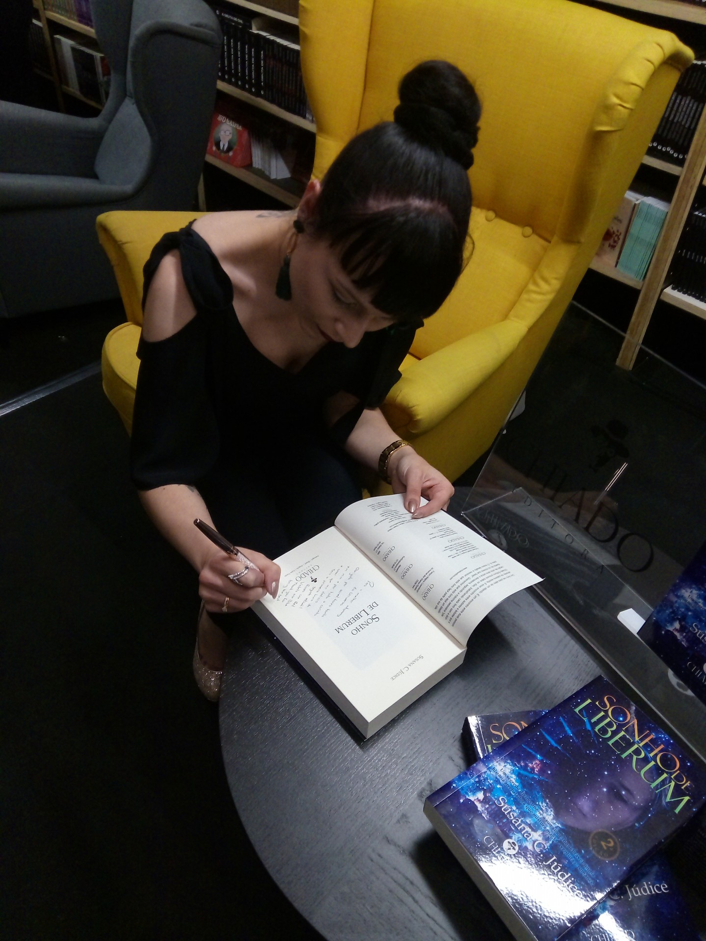 Mulher assinando um livro numa cadeira amarela junto a mesa com outro livro