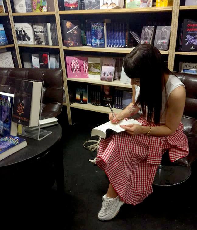 Mulher sentada a autografar livro em livraria, com saia xadrez vermelha e branca e tênis brancos