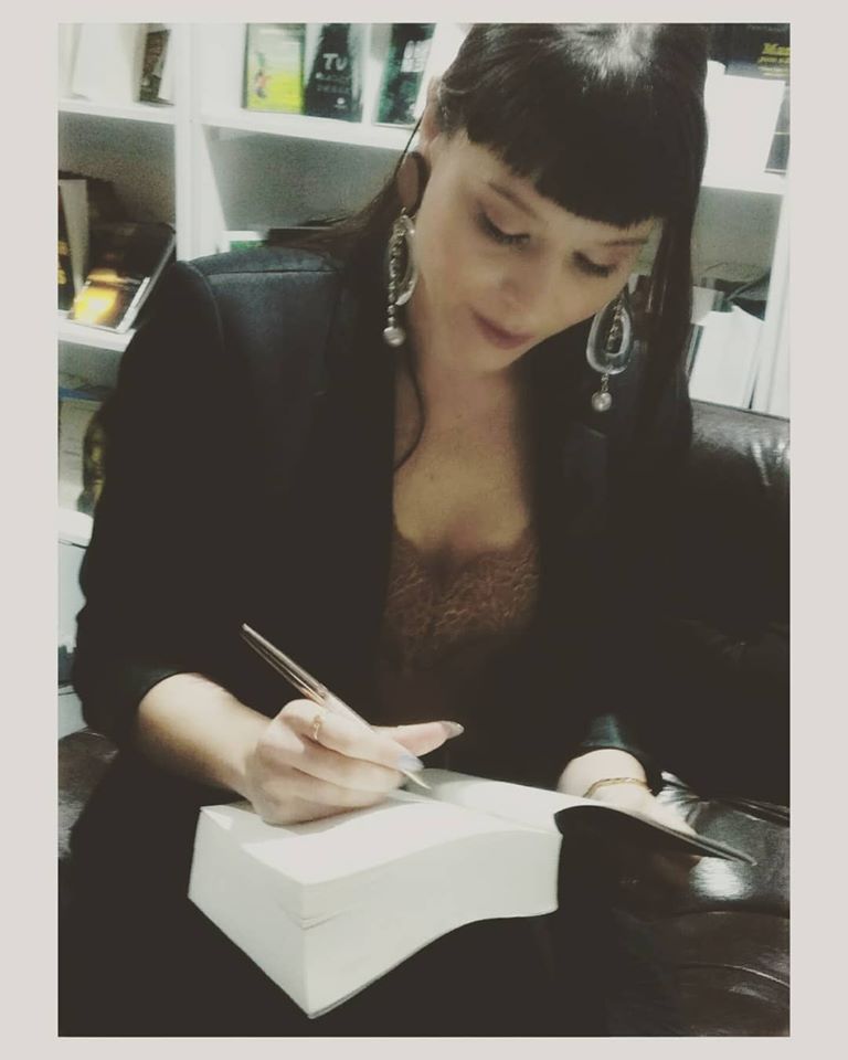 Mulher a assinar livro grosso com casaco preto e brincos grandes sentada num sofá escuro