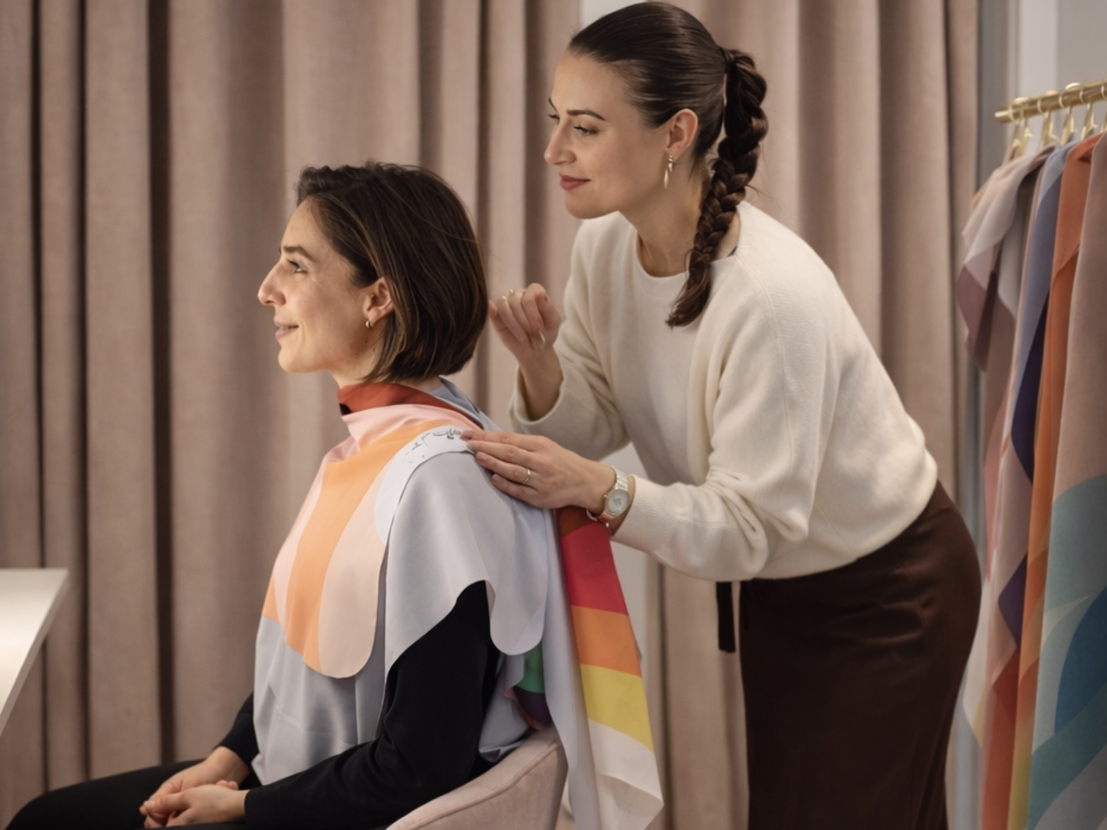 Duas mulheres, uma sentada com tecido colorido nos ombros e outra a ajudar, em frente a cortinas cor de rosa.