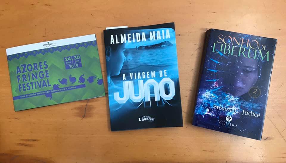 Três itens sobre uma mesa: panfleto do Azores Fringe Festival e dois livros intitulados 'A Viagem de Juno' e 'Sonhos Liberum'.