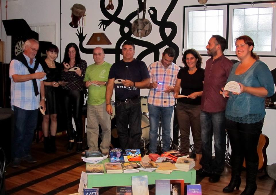 Grupo de oito pessoas em pé junto a uma mesa com livros e objetos em sala interior com arte na parede