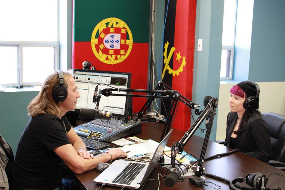 Estúdio de rádio com duas pessoas e bandeiras de Portugal e Angola ao fundo