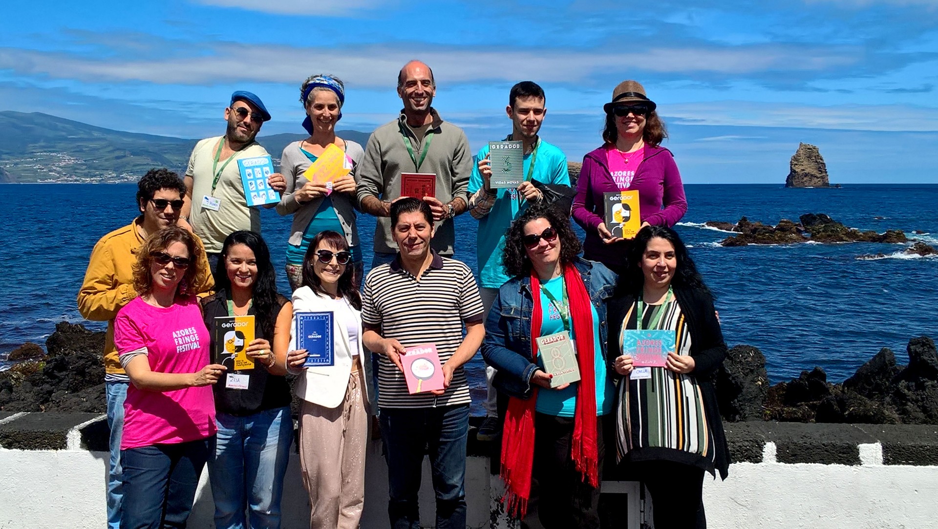 Grupo de pessoas segurando livros coloridos, junto ao mar com céu azul e nuvens.
