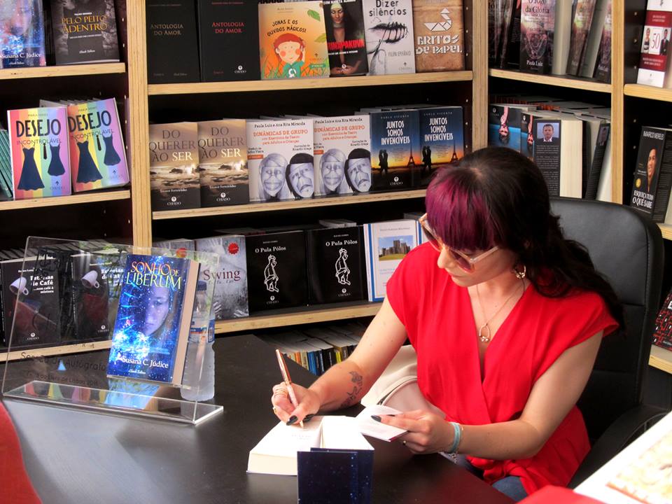 Mulher a autografar livro numa mesa com prateleiras de livros ao fundo