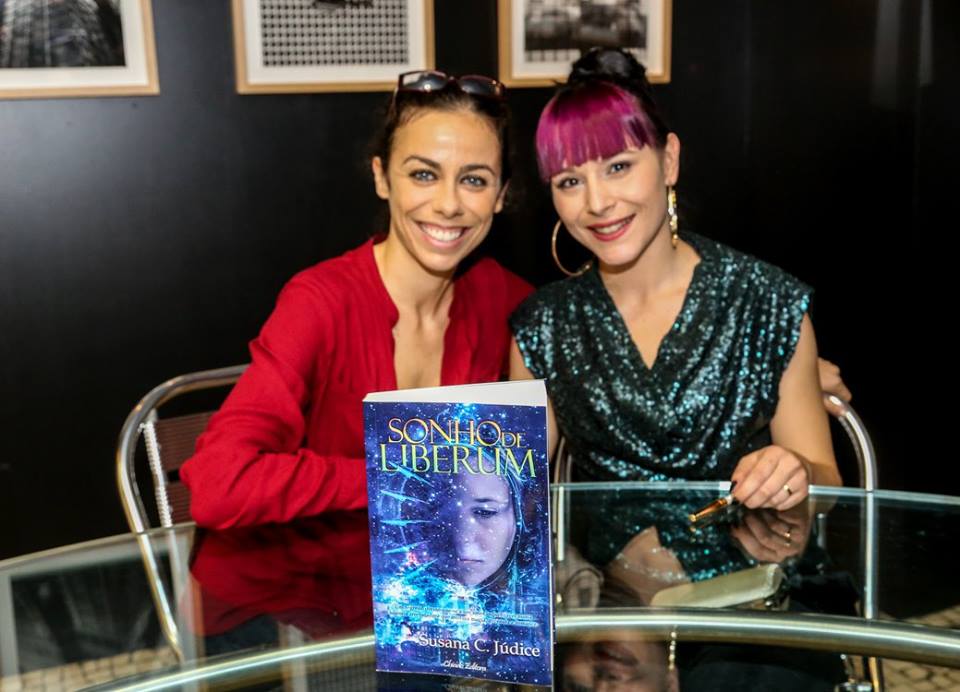 Duas mulheres sentadas atrás de uma mesa de vidro com o livro 'SONHO DE LIBERDADE' em destaque.
