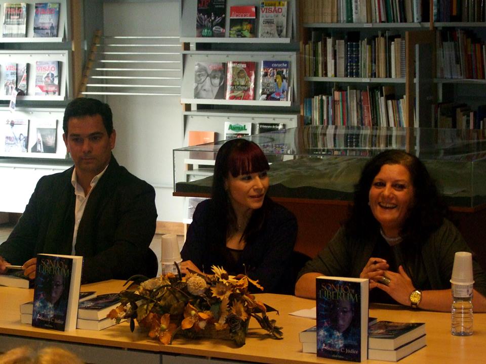 Três pessoas sentadas atrás de mesa com livros e flores numa biblioteca