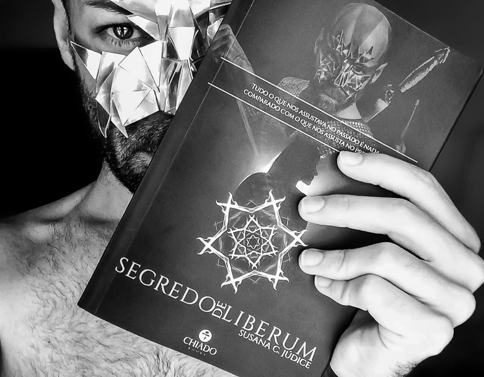 Homem com máscara prateada segurando livro SEGREDO LIBERUM