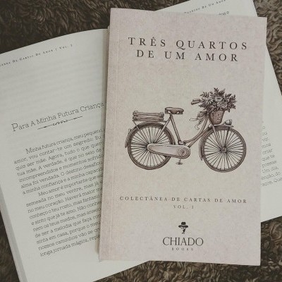 Capa do livro 'Três Quartos de Um Amor' com ilustração de bicicleta e livro aberto sobre tapete castanho.