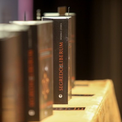 Livros alinhados numa prateleira, destaque para livro com título SEGREDOLIBERUM na lombada preta com texto laranja e branco