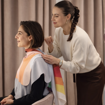 Duas mulheres, uma sentada com tecido colorido nos ombros e outra a ajudar, em frente a cortinas cor de rosa.