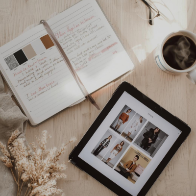 Mesa com tablet a mostrar moda, caderno aberto com anotações, óculos, caneca de chá e plantas secas