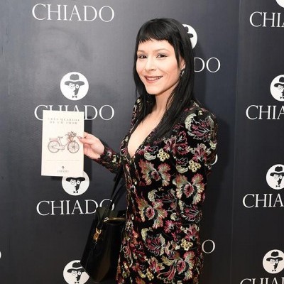 Mulher com vestido floral segurando livro e flor em frente a painel CHIADO