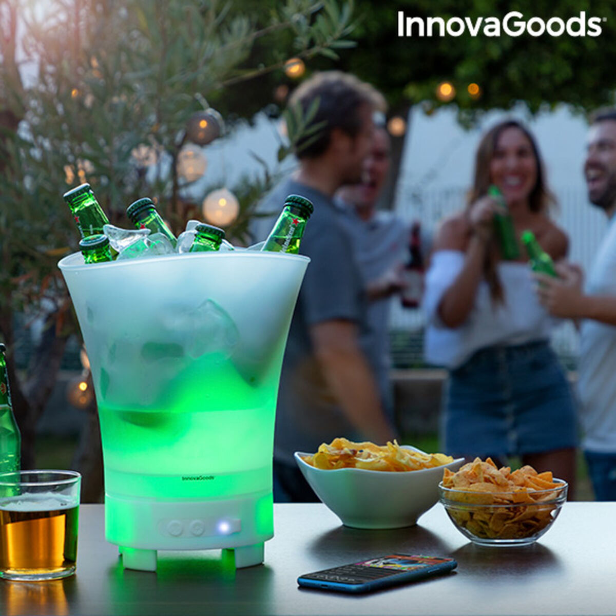 Balde de Gelo LED com Altifalante Recarregável Sonice InnovaGoods Balde de gelo com luz verde, garrafas e snacks numa mesa ao ar livre com pessoas ao fundo
