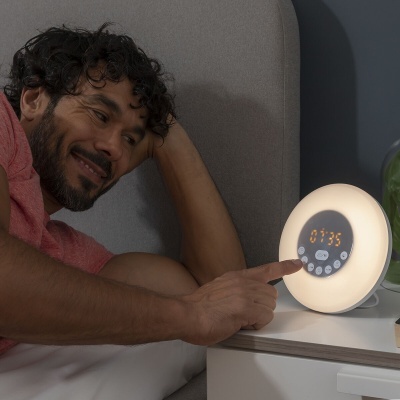 Relógio despertador digital redondo com ecrã e luz branco quente numa mesa de cabeceira ao lado da cama.