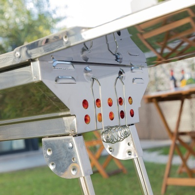 Churrasqueira metálica inoxidável com detalhes vermelhos e molas em jardim com móveis de madeira