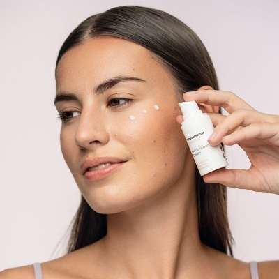 Mulher aplicando creme facial com frasco branco ao lado do rosto