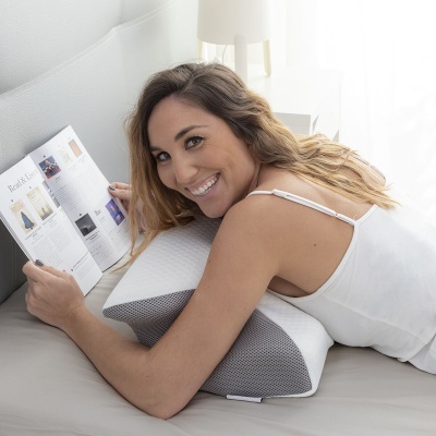 Mulher deitada sobre almofada de apoio branca e cinza, lendo revista na cama