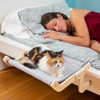 Cama suspensa para gato em madeira clara e tecido cinza ao lado de cama branca com mulher de pijama vermelho bordô dormindo