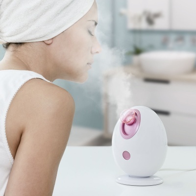Vaporizador facial branco com detalhes rosa a emitir vapor junto ao rosto de mulher com toalha na cabeça