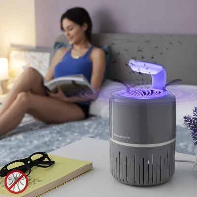 Dispositivo eletrônico cinzento com luz roxa num quarto, mulher a ler ao fundo, mesa com óculos e livro