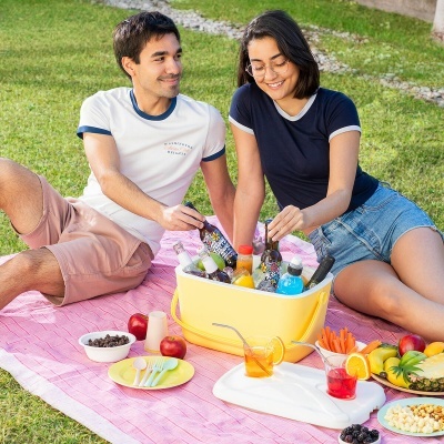 Casal fazendo piquenique com caixa térmica amarela, frutas e bebidas sobre manta rosa no relvado