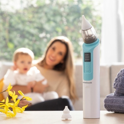 Aspirador nasal elétrico branco e azul claro numa mesa com acessórios e fundo desfocado de mulher e bebé