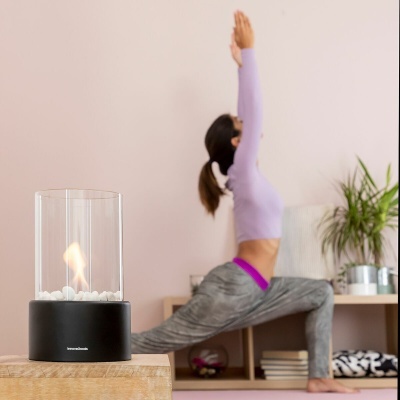 Lareira decorativa moderna com chama e pedras brancas numa mesa de madeira, senhora a fazer yoga ao fundo