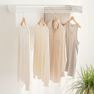 Roupas claras penduradas em cabides num varal branco de parede com toalhas e planta ao lado.