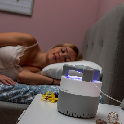 Dispositivo elétrico branco e cinzento com luz azul numa mesa ao lado de cama com mulher adormecida