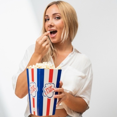 Mulher loira com camisa branca segurando balde de pipocas azul, branco e vermelho com texto 'POP CORN'