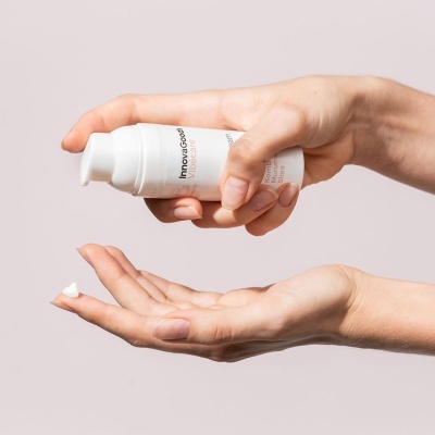 Frasco branco com creme sendo aplicado nas mãos contra fundo bege claro