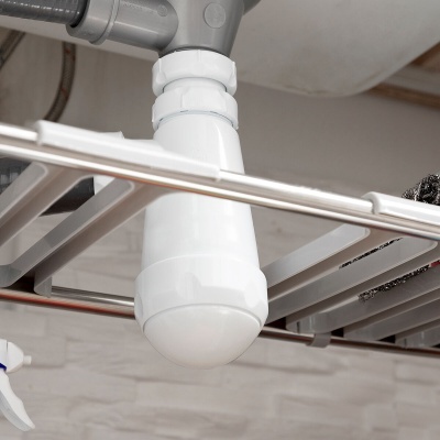 Sifão branco instalado sob pia com estrutura metálica e parede de azulejos brancos.