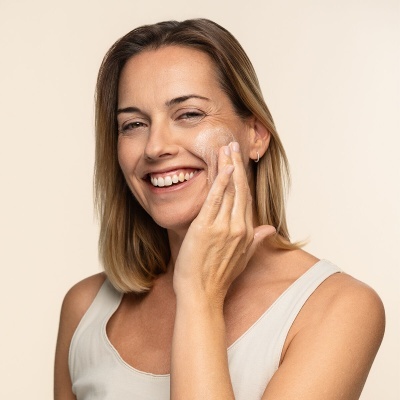 Mulher a aplicar creme facial branco na pele com top branco e fundo bege