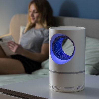 Dispositivo cilíndrico branco com anel azul luminoso numa mesa branca, mulher sentada num sofá atrás.