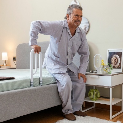Homem a usar barra de apoio branca fixada na cama em quarto com criado-mudo e quadro decorativo