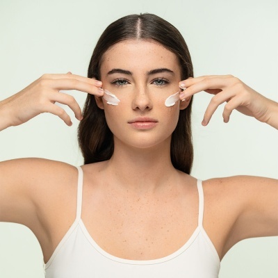 Mulher a aplicar creme facial em bochechas, vestindo camisola branca