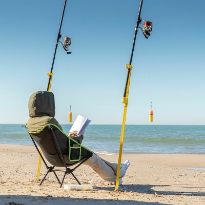 Pessoa sentada em cadeira de campismo cinza com detalhes verdes a ler livro na praia junto a varas de pesca amarelas.