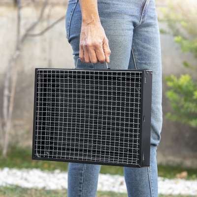 Grelhador portátil quadrado de metal preto com grelha metálica e pega, na mão de pessoa com calças de ganga azul ao ar livre
