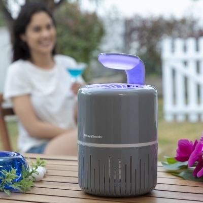 Dispositivo elétrico cinza escuro com cobertura roxa numa mesa de madeira, mulher desfocada ao fundo
