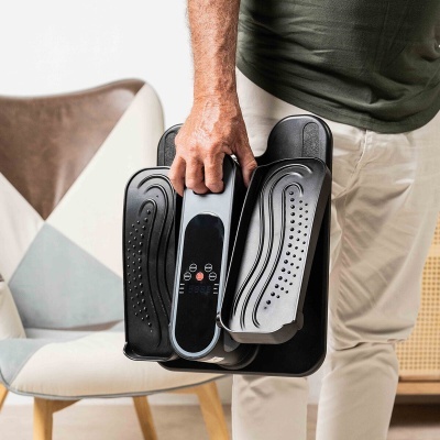 Dispositivo portátil de fitness preto com controles e suporta pés, segurado por uma mão.