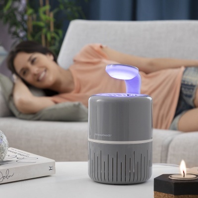 Purificador de ar cinzento com luz violeta sobre mesa branca com sofá e mulher ao fundo
