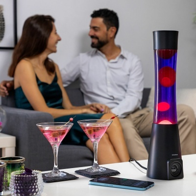 Lâmpada de lava azul com bolhas vermelhas numa mesa branca com cocktails, smartphone e velas, casal no sofá ao fundo.
