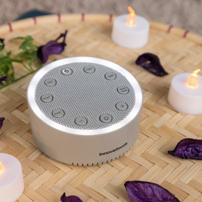 Coluna de som redonda prata com botões circulares e iluminação LED, sobre superfície de bambu com velas LED e folhas secas roxas