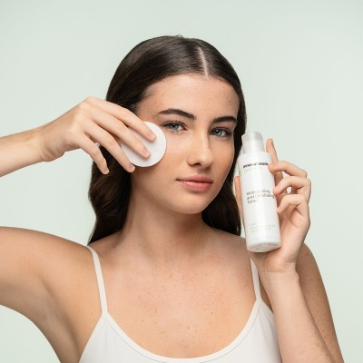 Mulher a usar almofada de algodão no rosto e a segurar frasco de produto cosmético branco