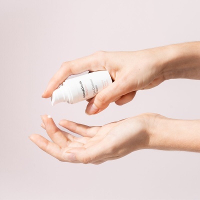 Frasco branco de creme sendo pressionado por uma mão, com texto parcial visível, em fundo claro