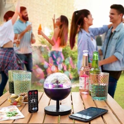 Mesa de madeira com copos, bebidas, dispositivo de luzes coloridas, comando remoto e smartphone, com pessoas ao fundo numa festa ao ar livre