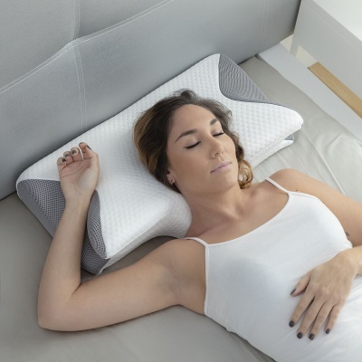 Almofada ortopédica branca e cinza numa cama com uma mulher a dormir de costas