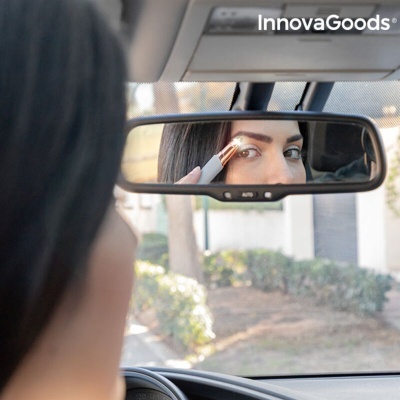 Pessoa no carro usando dispositivo elétrico branco e dourado para sobrancelhas refletido em espelho retrovisor.