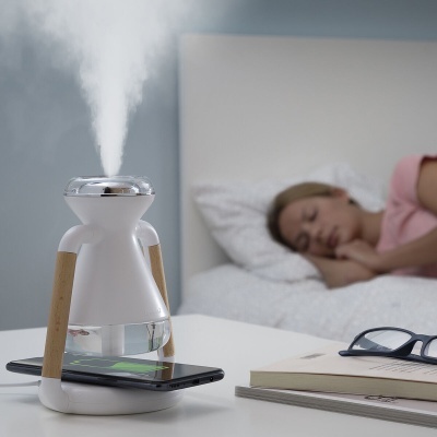 Humidificador de ar branco a emitir vapor numa mesa com smartphone e livros, mulher a dormir ao fundo